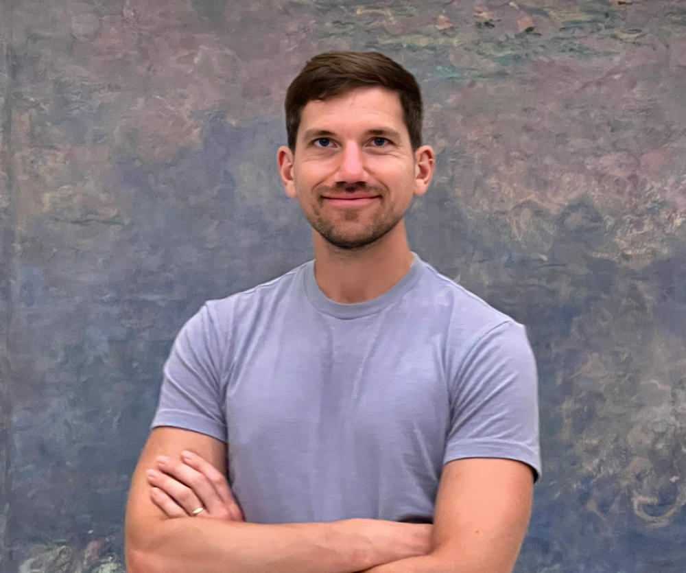 Photograph of Tim Gorichanaz, a man in his mid-30s with short brown hair and some stubble, with his arms folded and a slight smile. He is wearing a lavender-colored t-shirt and standing in front of an abstract Impressionist painting, one of Monet's monumental water lilie paintings at the Orangerie Museum in Paris.